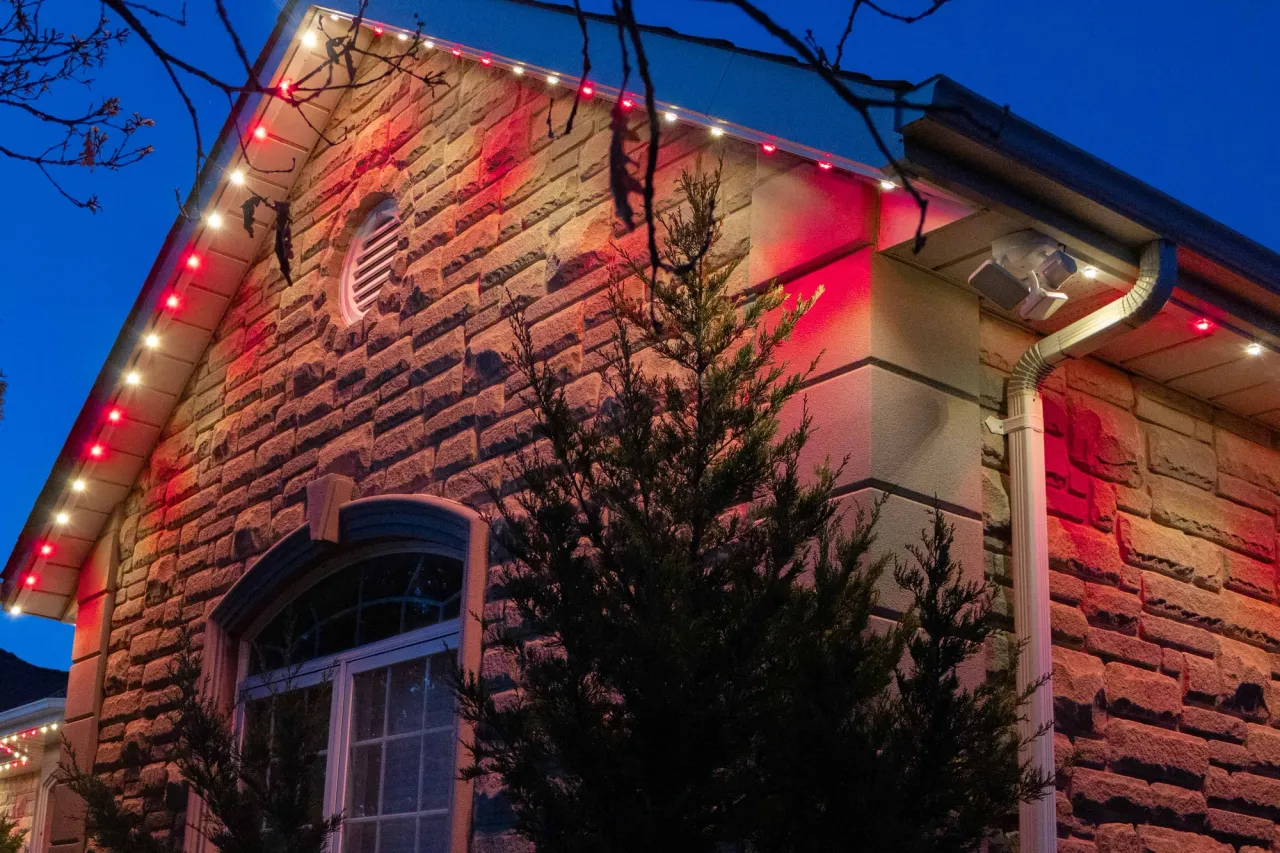 A home with a stone facade and round-topped window with Gemstone lights installed along the eaves, lit up in red and white lights at dusk.