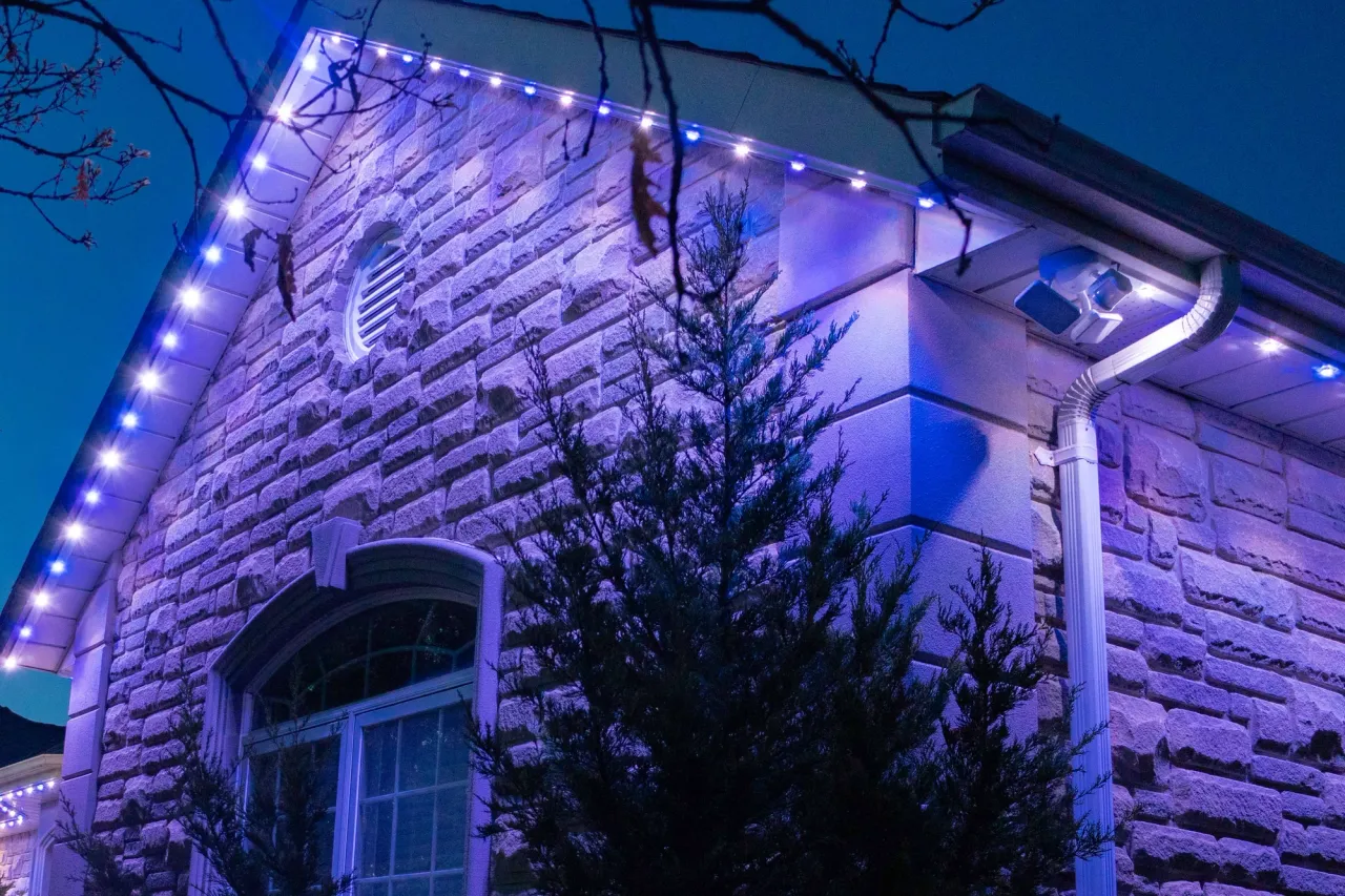 A home with a stone facade and round-topped window with Gemstone lights installed along the eaves, lit up in blue/purple lights at dusk.