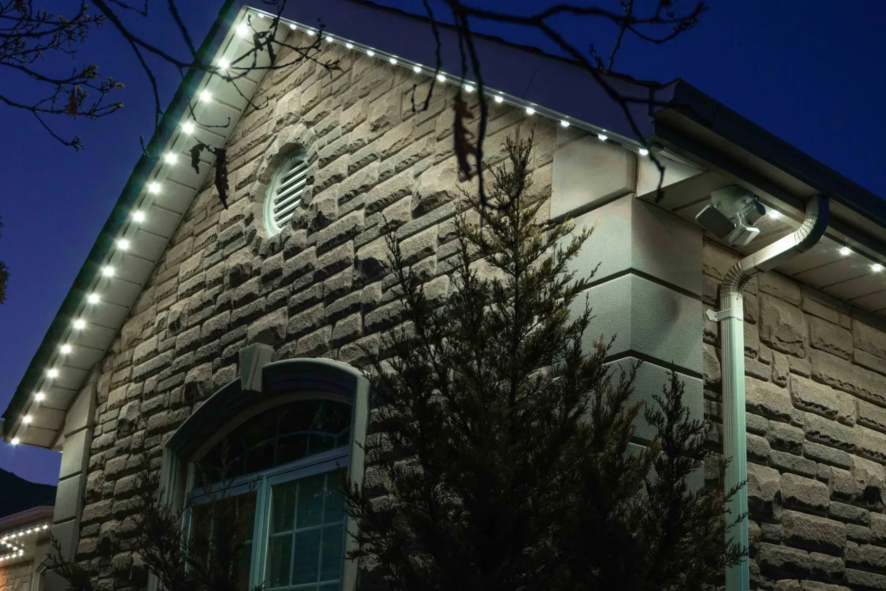 A home with a stone facade and round-topped window with Gemstone lights installed along the eaves, lit up in white lights at dusk.