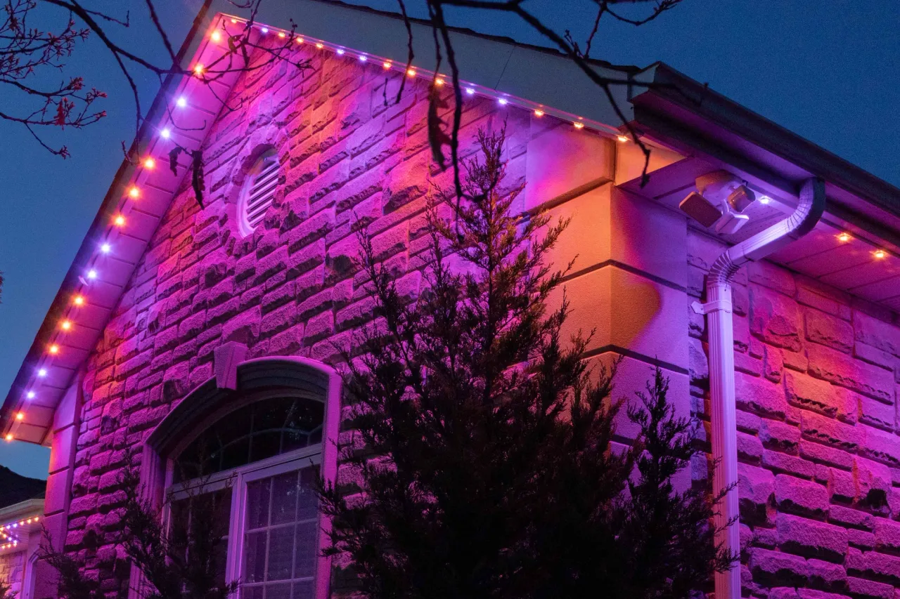 A home with a stone facade and round-topped window with Gemstone lights installed along the eaves, lit up in orange and purple lights at dusk.